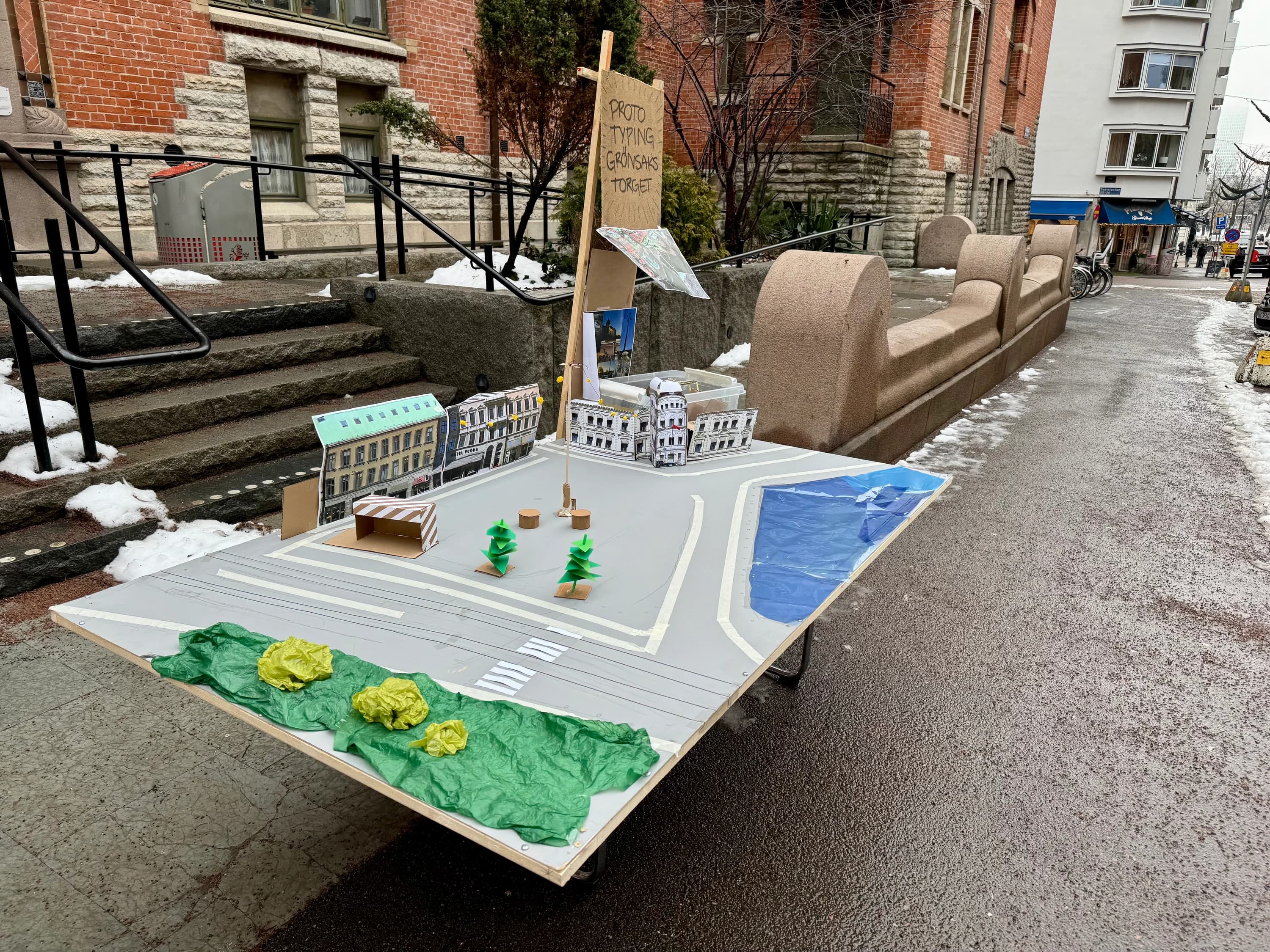The model outdoors on a snowy street outside HDK-Valand, with a hand-lettered cardboard sign reading Prototyping Grönsakstorget.