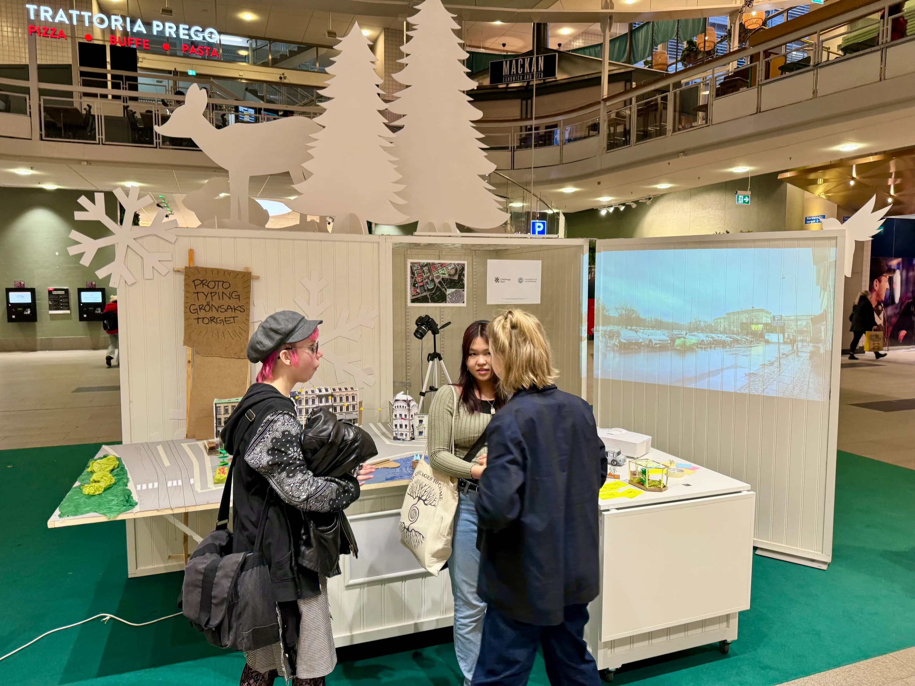 The stall set up inside the Nordstan shopping centre, three people standing around the model in conversation, a projection of the real square behind it.