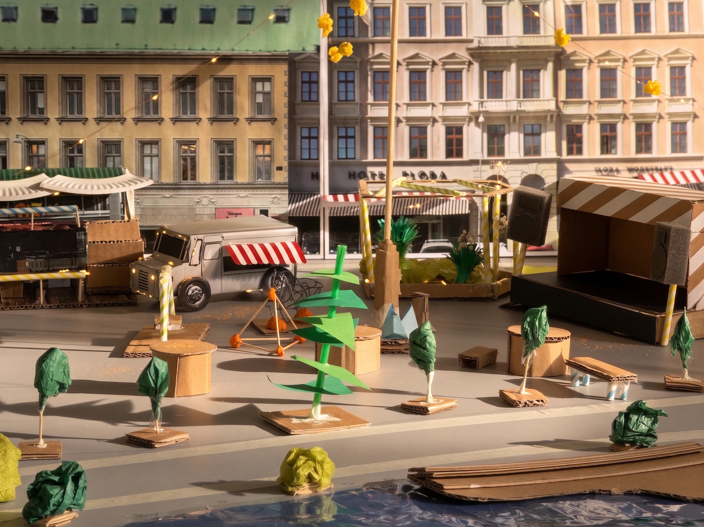 Close view of the populated model: cardboard market stalls under striped awnings, a delivery van, paper trees, benches and string lights against printed facades.